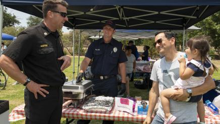 Assemblymember Rendon holding his daughter having a conversation with first responders by a tent labelled with the Assemblymember's logo