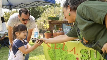 Assemblymember Rendon stands behind his daughter while she accepts a succulent from someone who is working a succulent table